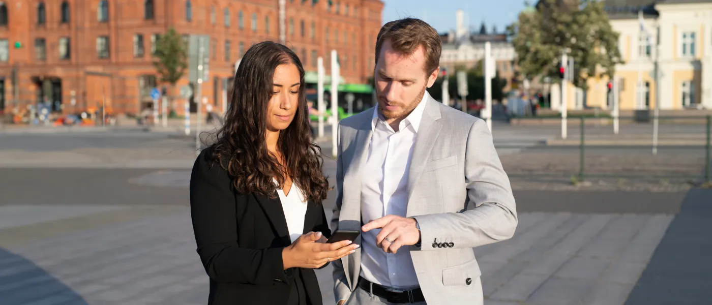 A woman and man standing in businss clothes outside. The woman is holding a mobile in her hand, the man are pointing at the screen. 
