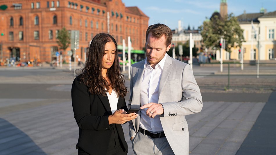 A woman and a man standing out side wearing business clothes. The woman in holding a mobile, the man in pointing at the screen. 