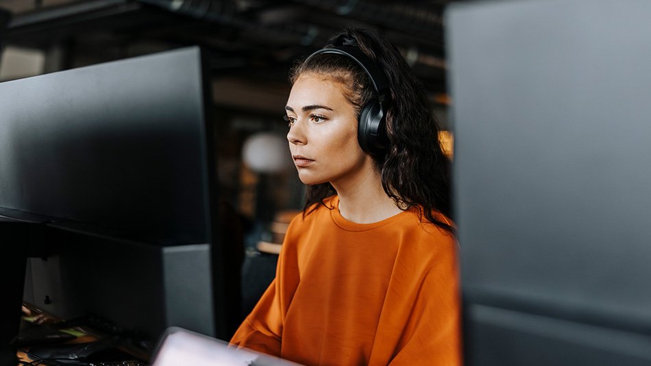 Girl sitting infront of a computer monitor working. She is wearing headphones.